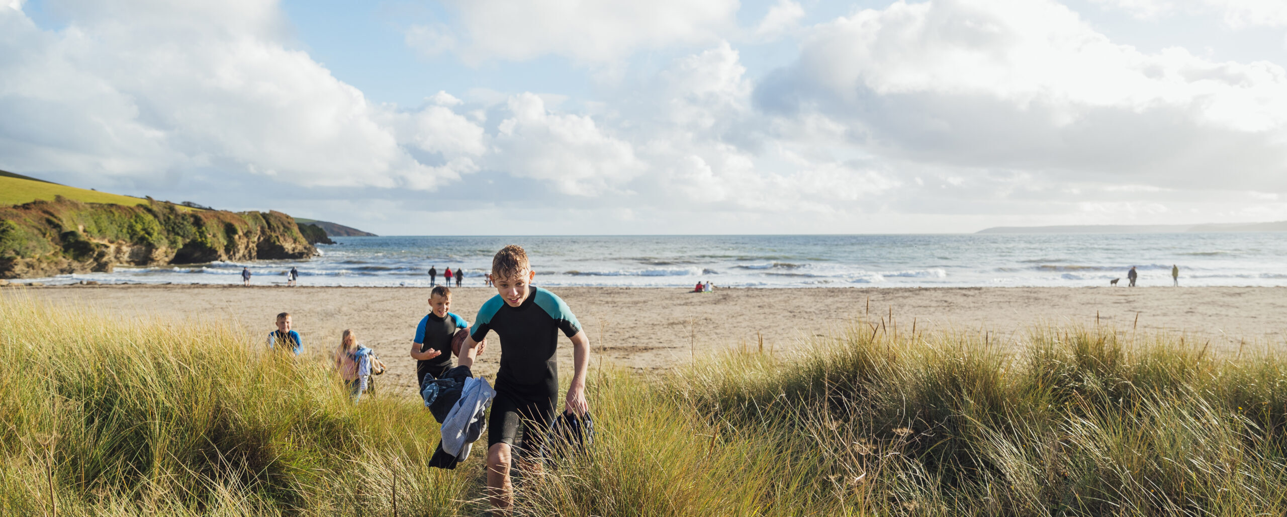 Boys earring wetsuits walking from the beach on a sunny day in autumn. They are on a staycation with their family to lower their carbon footprint and support the local economy.